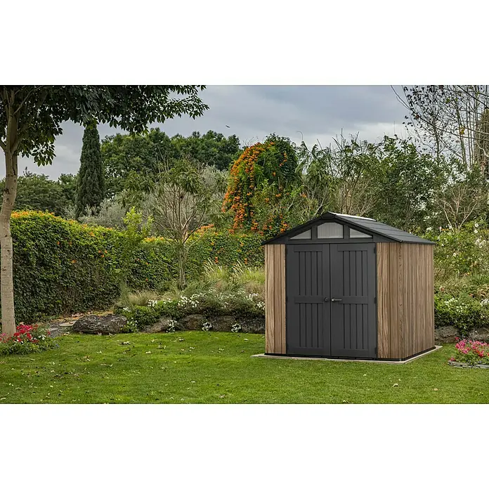 Keter Gerätehaus Stronghold 8x8 - Außenmaß inkl. Dachüberstand (B x T): 248,5 x 248,5 cm, Kunststoff, Eiche/Schwarz - Ein geräumiges Gartenhaus mit Doppeltür und Holzverkleidung vor üppigem Grün.