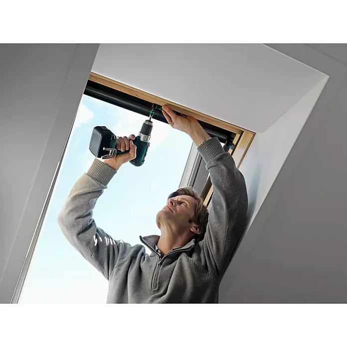 Velux Solar-Nachrüst-Set KSX 100 - Passend für: VELUX Schwingfenster GGU/GGL 102 – 810/C02 – U10, VL Y und VU Y - Ein Mann befestigt mit einem Akkuschrauber ein VELUX-Dachfenster. Das Fenster erhellt den Raum mit natürlichem Licht und sorgt für frische Luft.