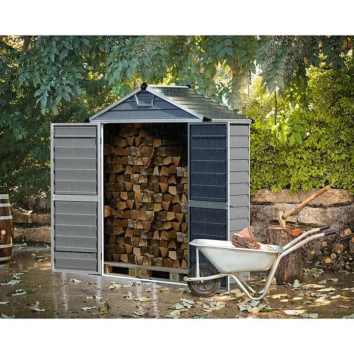 Palram – Canopia Gartenhaus Skylight - Außenmaß inkl. Dachüberstand (B x T): 185 x 90 cm, Kunststoff, Anthrazit/Grau - Ein graues Gartenhaus mit geöffneten Türen, gefüllt mit Holz, steht im Regen. Eine Schubkarre und ein Hackklotz stehen davor.