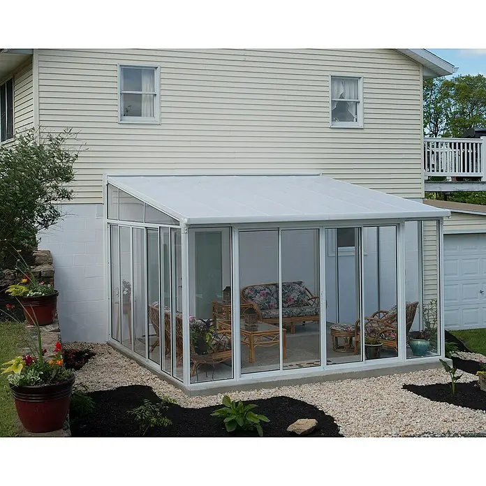 Palram – Canopia Porche acristalado San Remo - An x Pr x Al: 4,25 x 4 x 3,1 m, Blanco tráfico - Un solárium con marcos blancos y paredes de vidrio perfectamente unido a una casa con revestimiento beige claro, creando una acogedora expansión.