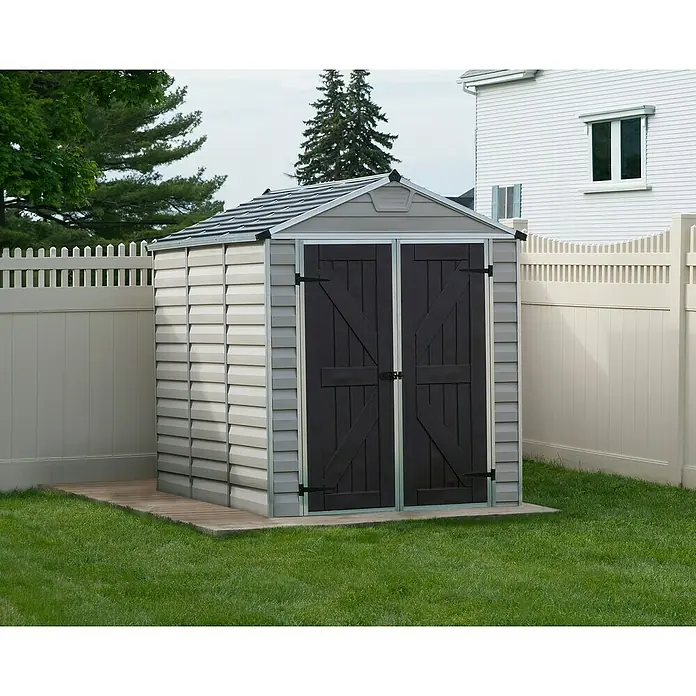 Palram – Canopia Gartenhaus Skylight 6x8 Tan - Außenmaß inkl. Dachüberstand (B x T): 185 x 229 cm, Kunststoff, Beige/Braun - Ein graues Kunststoff-Gartenhaus mit Doppeltüren auf einer Holzplattform, umgeben von einem grünen Rasen und einem weißen Zaun.