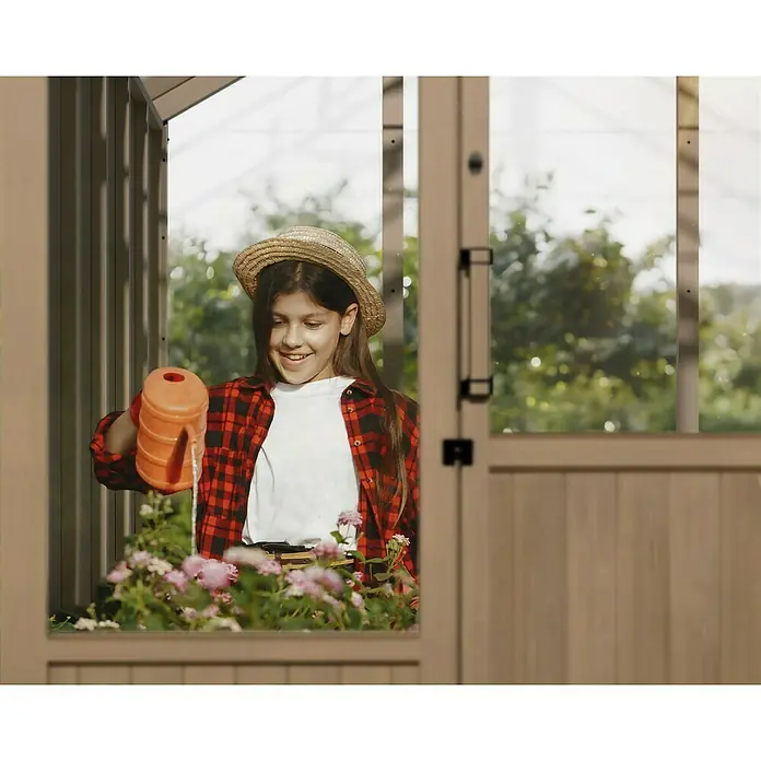 Palram – Canopia Invernadero Natura - Medidas ext. (An x Pr): 249 x 356 cm, Plástico, 4 mm, Natural - Una joven está regando flores que crecen en un contenedor con una regadera naranja dentro de un invernadero.