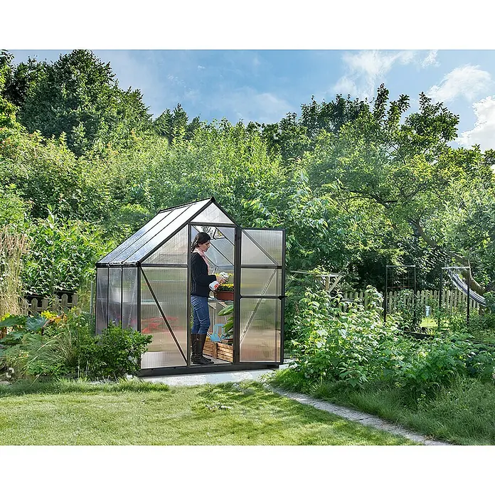 Palram – Canopia Invernadero Mythos - Medidas ext. (An x Pr): 185 x 186 cm, Material de la pared lateral: Plástico, 4 mm, Gris - Un invernadero con una mujer dentro cuidando las plantas. El invernadero se encuentra en una zona de jardín con vegetación alrededor.
