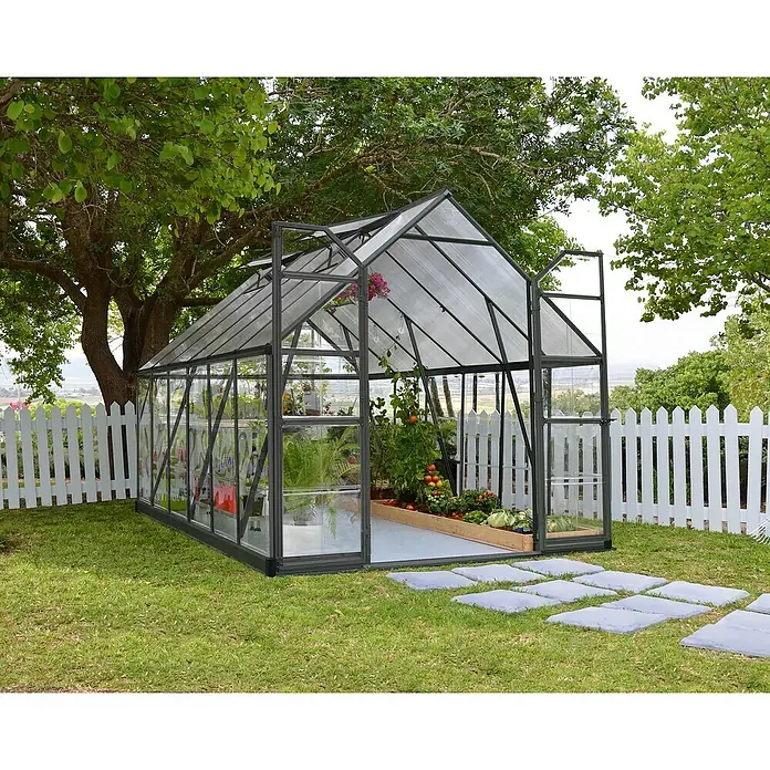 Palram – Canopia Invernadero Balance - Medidas ext. (An x Pr): 244 x 367 cm, 0,7 mm, Gris - Un invernadero espacioso con plantas dentro está situado en un jardín, rodeado por una valla blanca y árboles.