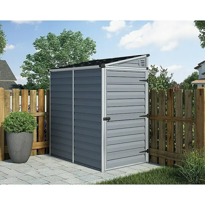 Palram – Canopia Caseta de jardín Skylight - Medidas ext. (An x Pr): 117 x 177 cm, Plástico, Antracita - Un cobertizo de jardín de plástico gris está ubicado sobre una superficie pavimentada, rodeado por una valla de madera y vegetación.