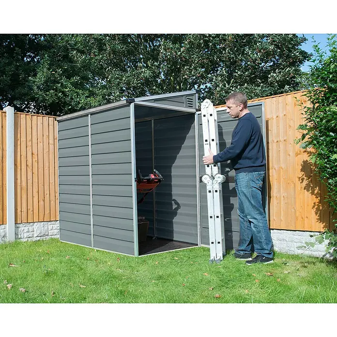 Palram – Canopia Caseta de jardín Skylight - Medidas ext. (An x Pr): 117 x 177 cm, Plástico, Antracita - Un hombre está metiendo una escalera de aluminio en un cobertizo de jardín gris, situado en un césped verde.