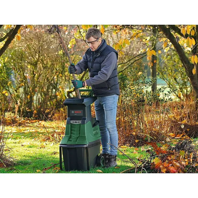 Bosch Elektro-Häcksler AXT 25 TC - Max. Aststärke: 4,5 cm, 2.500 W - Ein Mann benutzt einen Bosch Gartenhäcksler, um Äste zu zerkleinern. Der Häcksler steht auf einer Wiese mit Herbstlaub.