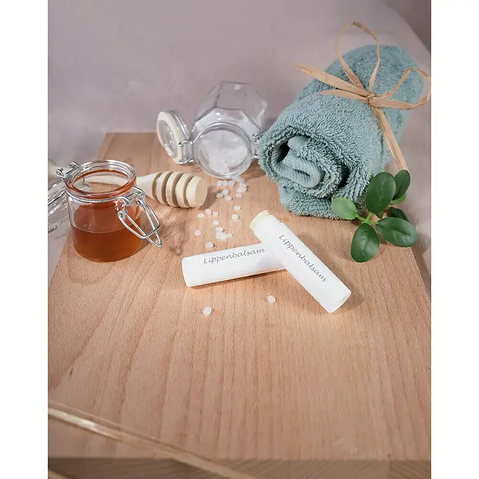 Glorex Bastel-Set Naturkosmetik Lippenbalsam - 14 -tlg. - Nahaufnahme von zwei Lippenbalsamstiften auf einem Holzbrett mit Honigglas, Wachspastillen, Spender und blauem Handtuch.