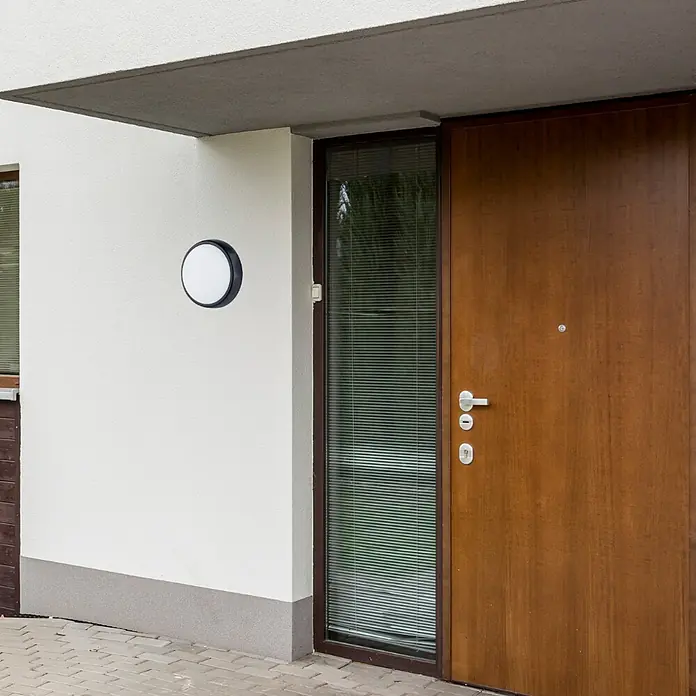 Luceco Aplique exterior LED Mini Circular - 5,5 W, 16 x 7 x 16 cm, Negro, Blanco, IP54 - Una luz redonda de exterior con marco negro está montada en una pared blanca, junto a una puerta de madera. Esta lámpara proporciona una iluminación exterior elegante.