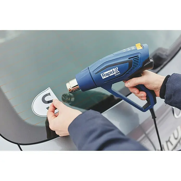 Rapid Heißluftgebläse R2000 - 2.000 W, Temperaturbereich: 60 °C - 600 °C - Ein Rapid R2000 Heißluftfön wird verwendet, um einen Aufkleber von einer Autoscheibe zu entfernen, ideal für Werkstattarbeiten.
