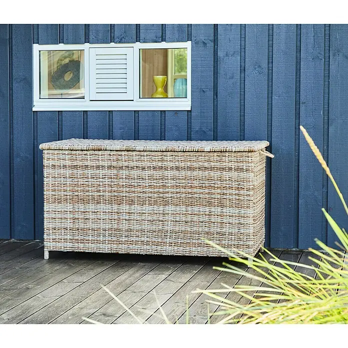 Baúl para jardín Gotland - 150 x 65 x 68 cm, Plástico, Arena - Una caja de almacenamiento de mimbre para exteriores. La cesta es de ratán claro y está sobre una terraza de madera frente a una pared de madera azul.