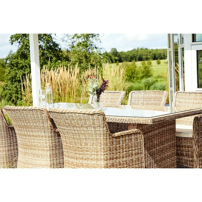 Sillón de jardín Gotland - An x Pr x Al: 79 x 63 x 99 cm, Arena - Primer plano de una mesa de ratán con tapa de cristal y sillas de ratán en un patio con vistas a la vegetación.