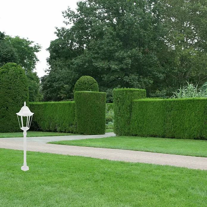 Panaled Vagg Lámpara de pie para exterior - Color del marco: Blanco, Sin bombillas - Una farola de jardín blanca sobre un césped bien cuidado, rodeada de setos y árboles verdes y exuberantes.