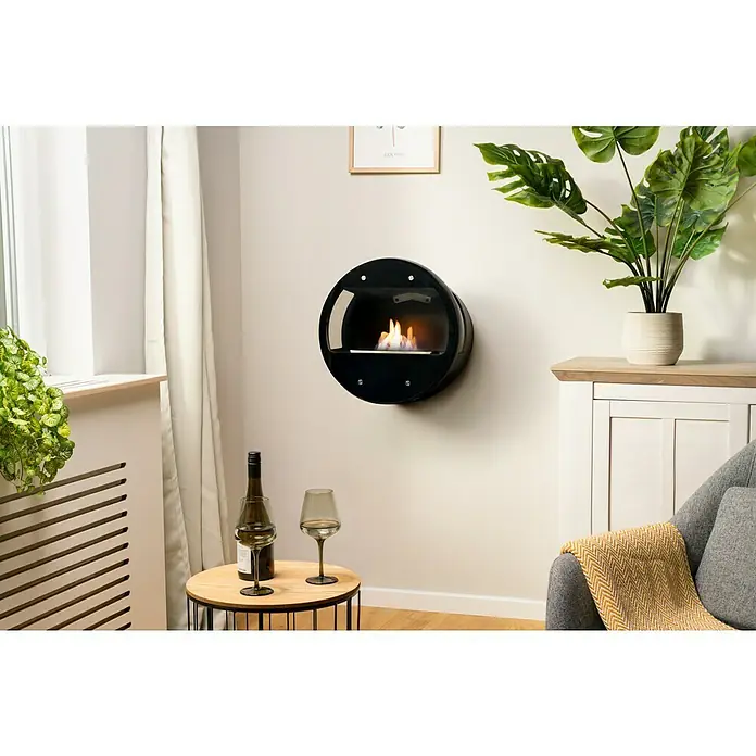 Aduro Bioethanol-Kamin B1 - 46 x 63 x 70 cm, Schwarz - Ein runder, schwarzer Wandkamin sorgt für Wärme und Atmosphäre. Daneben stehen Weinflasche und Gläser auf einem Beistelltisch.