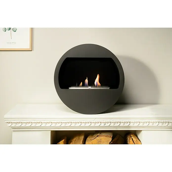 Aduro Bioethanol-Kamin B2 - 46 x 63 x 70 cm, Schwarz - Runder Bioethanol-Kamin in Grau auf einem weißen Kaminsims mit Holzscheiten, der eine warme und moderne Atmosphäre schafft.