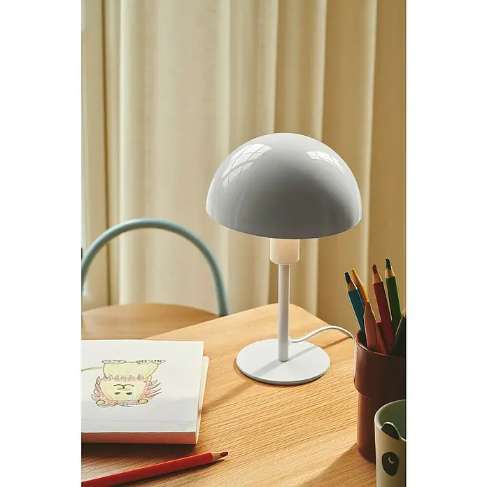 Nordlux Lámpara de sobremesa Ellen mini - 40 W, L x An x Al: 16 x 16 x 25 cm, Blanco, E14 - Primer plano de una lámpara de mesa blanca con pantalla hemisférica sobre una mesa de madera con lápices de colores y un dibujo.