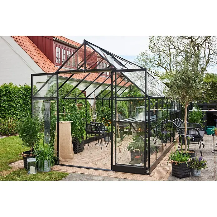 Halls Qube Gewächshaus 816 - Außenmaß inkl. Dachüberstand (B x T): 261 x 508 cm, 3 mm, Schwarz - Ein Gewächshaus mit schwarzem Rahmen und Glas im Garten, gefüllt mit Topfpflanzen, Sitzgelegenheiten und kleinen Orangenbäumen, als gemütlicher Gartenzimmer.