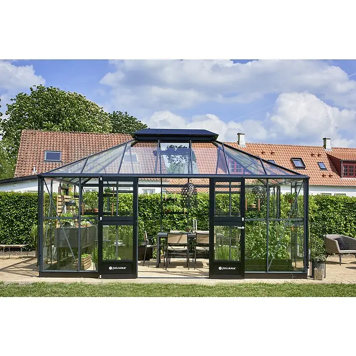 Juliana Gewächshaus Grand Oase - Außenmaß inkl. Dachüberstand (B x T): 368 x 511 cm, 3 mm, Anthrazit/Schwarz - Das Bild zeigt ein Juliana Gewächshaus mit transparenten Wänden, das Pflanzen beherbergt und einen Essbereich mit Tisch und Stühlen bietet.