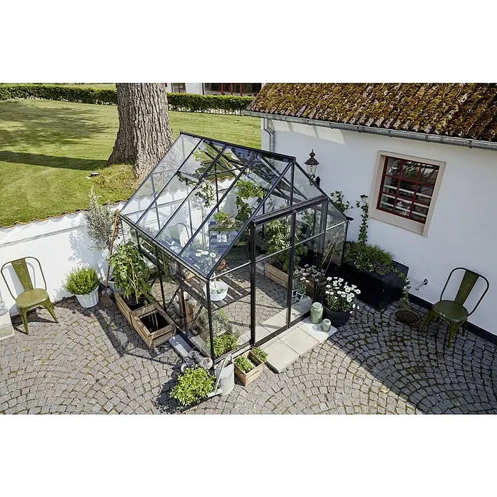 Halls Gewächshaus Qube 88 - Außenmaß inkl. Dachüberstand (B x T): 261 x 261 cm, 3 mm, Schwarz - Ein Gewächshaus mit Pflanzen und Kräutern in einem Garten. Der Außenbereich ist mit Stühlen und Pflastersteinen gestaltet.