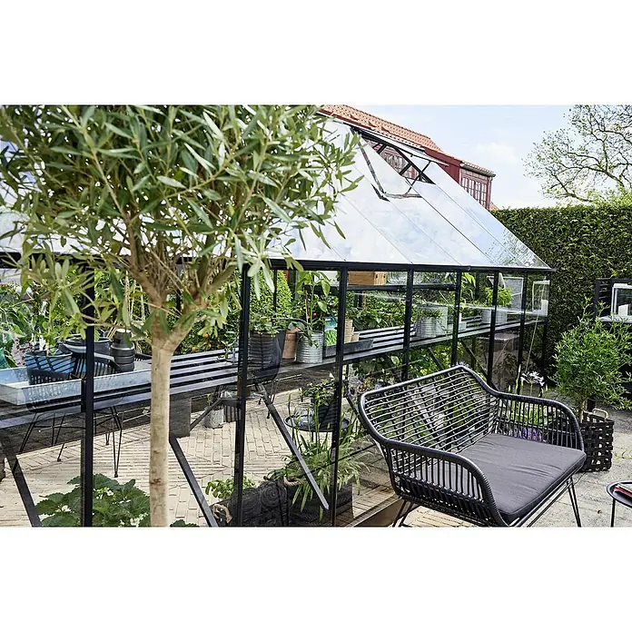 Halls Gewächshaus-Tisch - Passend für: Juliana Gewächshaus Qube 816 - Ein modernes Gewächshaus mit schwarzem Rahmen und Glaswänden, umgeben von üppigem Grün und einer stilvollen Sitzgelegenheit.