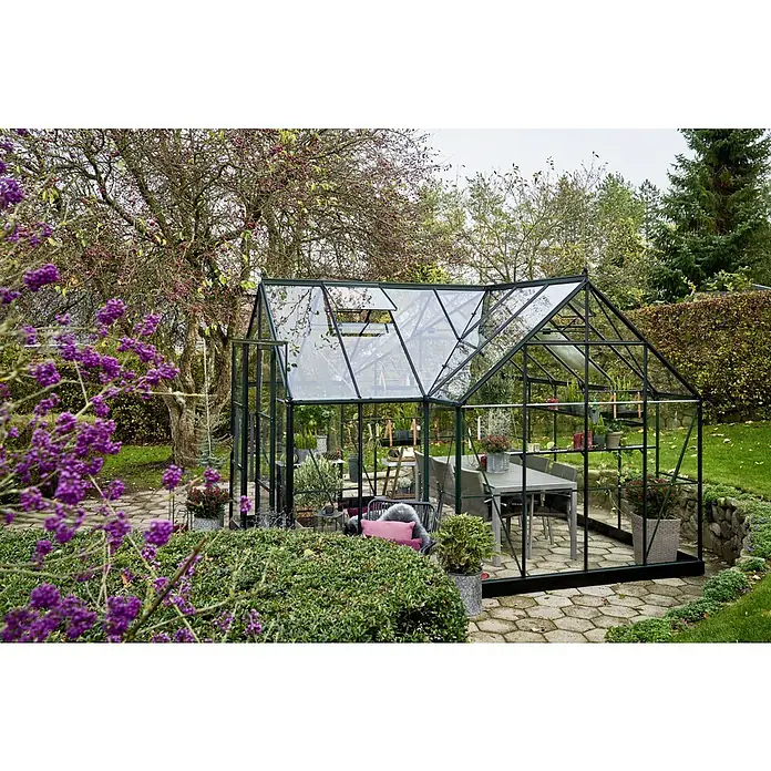 Halls Gewächshaus Garden Room - Außenmaß inkl. Dachüberstand (B x T): 389 x 390 cm, Farbe Gestell: Grün, Wandstärke: 3 mm - Ein elegantes Gewächshaus mit dunklem Rahmen fügt sich harmonisch in einen Garten mit blühenden Pflanzen und einem einladenden Esstisch im Inneren ein.