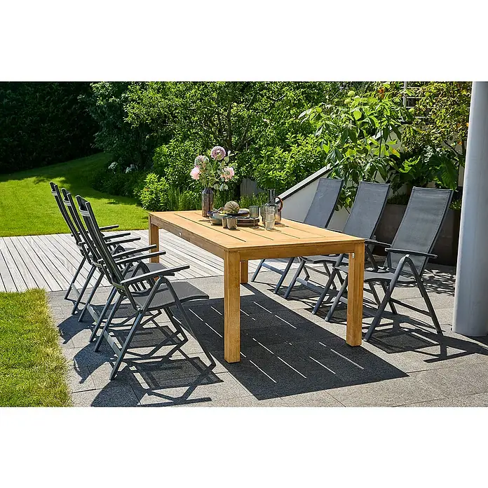 Sunfun Gartenmöbel-Set Laura/Maja - 7 -tlg., Akazie, Aluminium, Textilen, Naturbraun/Night-Grey - Outdoor-Szene mit einem Gartentisch aus Holz und sechs grauen Klappstühlen, umgeben von üppigem Grün und hellen Steinfliesen für sommerliche Entspannung.