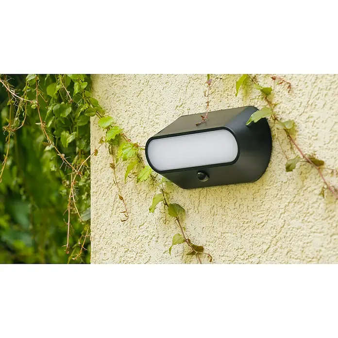 Lavida Solar-Außenwandleuchte - 5 W, L x B x H: 25 x 12 x 6,5 cm, Schwarz, IP54, 500 lm - Eine moderne, schwarze Wandleuchte mit Bewegungssensor, umgeben von Kletterpflanzen, die eine stilvolle Außenbeleuchtung bietet.