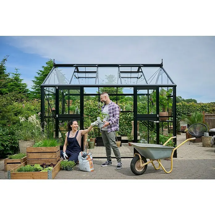 Juliana Gewächshaus Jubii 60 - Außenmaß inkl. Dachüberstand (B x T): 368 x 296 cm, Material Seitenwand: Glas, 3 mm, Schwarz - Ein Gewächshaus mit einem Paar, das gärtnert, mit Holzkisten, einer Schubkarre und viel Grün. Ideal für Gartenliebhaber.