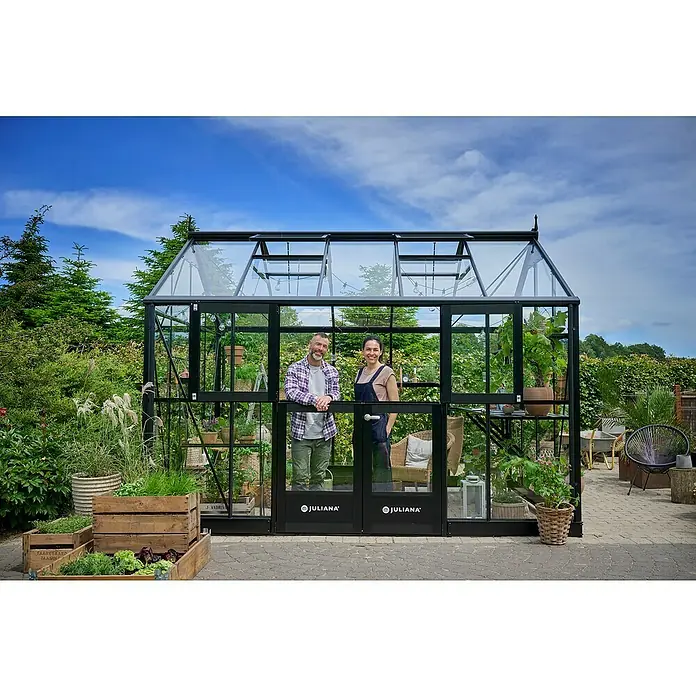 Juliana Gewächshaus Jubii 60 - Außenmaß inkl. Dachüberstand (B x T): 368 x 296 cm, Material Seitenwand: Glas, 3 mm, Schwarz - Ein großes Juliana Gewächshaus mit zwei Personen davor, eingebettet in eine grüne Gartenlandschaft unter blauem Himmel.