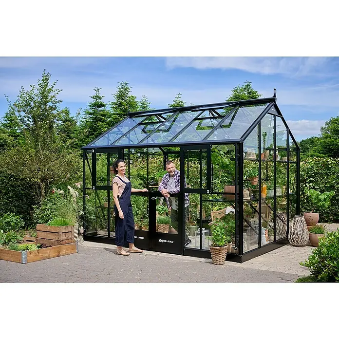 Juliana Gewächshaus Jubii 60 - Außenmaß inkl. Dachüberstand (B x T): 368 x 296 cm, Material Seitenwand: Glas, 3 mm, Schwarz - Ein Juliana Gewächshaus mit zwei Personen davor, umgeben von üppigem Grün. Perfekt für Hobbygärtner und zur stilvollen Pflanzenanzucht.