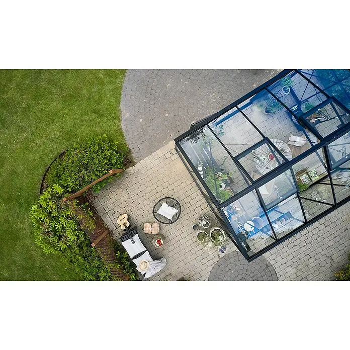 Juliana Gewächshaus Jubii 60 - Außenmaß inkl. Dachüberstand (B x T): 368 x 296 cm, Material Seitenwand: Glas, 3 mm, Schwarz - Ein Gewächshaus aus Glas und Metall, umgeben von einem Garten mit Pflanzen und Möbeln, lädt zum Entspannen und Genießen ein.