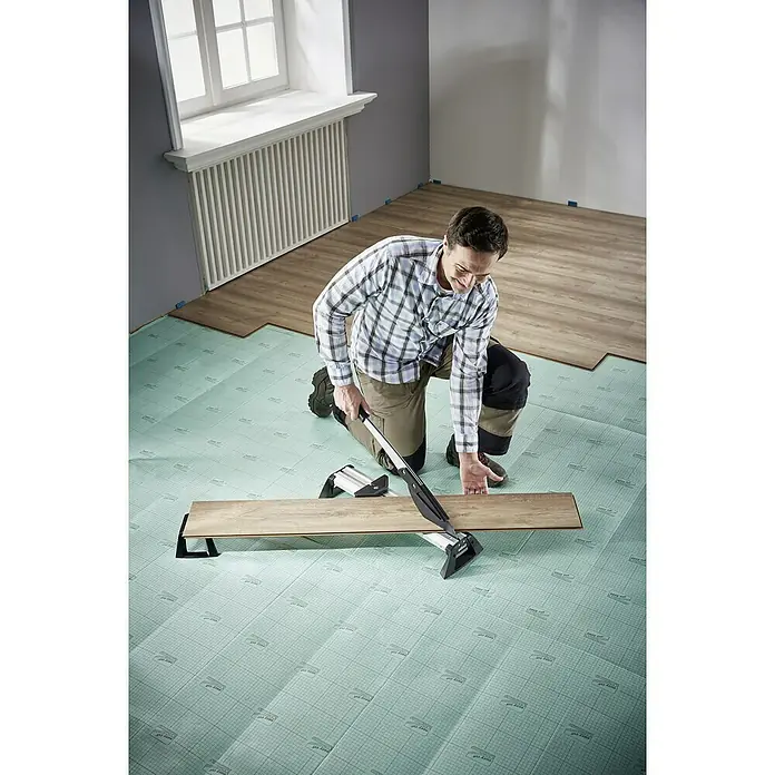 Wolfcraft Cortaláminas de parquet VLC 800 - Ancho de corte: 465 mm, Aluminio - Un hombre está arrodillado cortando un trozo de suelo laminado con un cortador de laminado. La capa inferior de color claro está impresa con una cuadrícula.