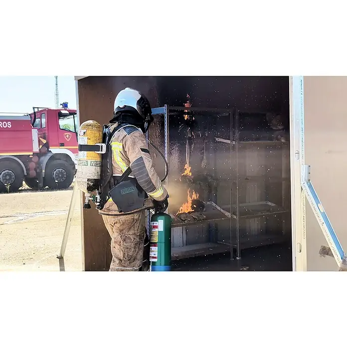 Simonrack Ecofire Extintor de incendios Fighting con gancho - 6 l - Un bombero con extintor combate un incendio en una sala de formación. Un camión de bomberos rojo espera al fondo como apoyo.