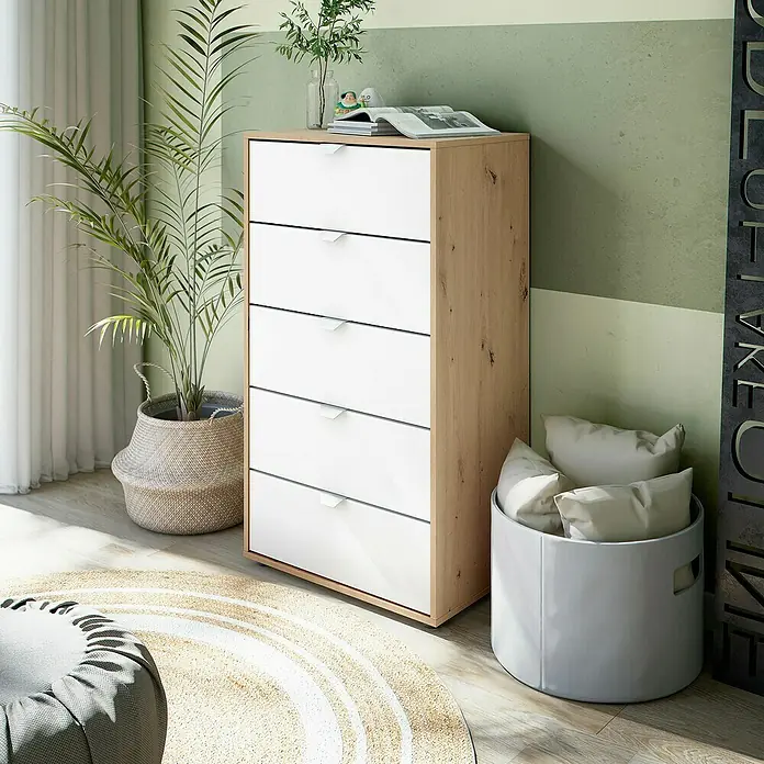 Cómoda Garona - L x An x Al: 40 x 60 x 109 cm, Roble Nodi/Blanco Artik, Número de cajones: 5 ud. - Cómoda con cinco cajones en blanco y aspecto de madera, junto a una planta de interior y una cesta de cojines.