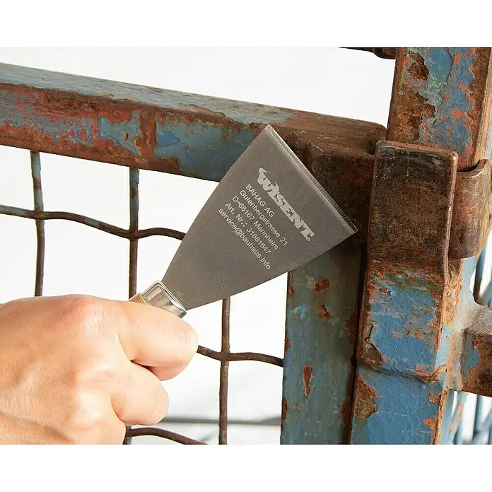 Wisent Entrosterspachtel - Klingenbreite: 60 mm, Länge: 180 mm - Nahaufnahme eines Wisent Spachtels beim Entfernen von Farbe von einem verrosteten Metalltor.