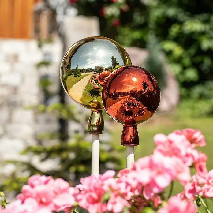 Windhager Glaskugel - Durchmesser: 12 cm, Rot - Zwei glänzende Gartenkugeln in Gold und Kupfer, die die Umgebung reflektieren, stehen inmitten von rosa Geranien.