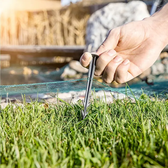 Windhager Laubschutznetz - L x B: 5 x 4 m, Kunststoff, Grün - Nahaufnahme einer Hand, die einen Hering zur Befestigung eines Netzes im Gras befestigt, ideal für Garten- und Landschaftsbauprojekte.
