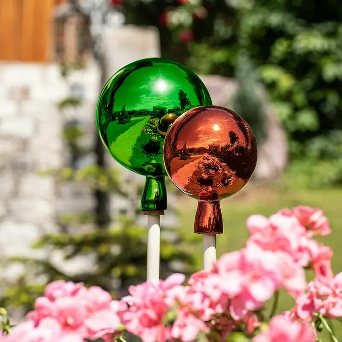 Windhager Glaskugel - Durchmesser: 12 cm, Orange - Nahaufnahme von zwei Gartenkugeln in Grün und Kupfer, die zwischen rosa Blumen im Garten stehen und Umgebungsdetails reflektieren.
