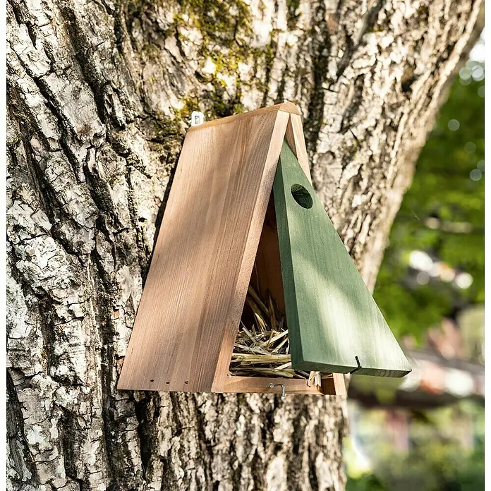 Windhager Nistkasten Delta - L x B x H: 14 x 18 x 28 cm, Birke, Braun/Grün - Ein dreieckiges Vogelhaus aus Holz in Natur und Grün hängt an einem Baum. Das Haus ist mit Stroh gefüllt und bietet einen sicheren Unterschlupf.