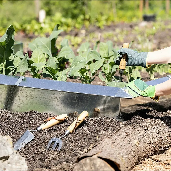 Windhager Schneckenblech Zaun-Element - Stahl, L x H: 115 x 25 cm - Gartenszene mit einer Person, die ein Metallbeet bearbeitet, mit Werkzeugen und Grünpflanzen.