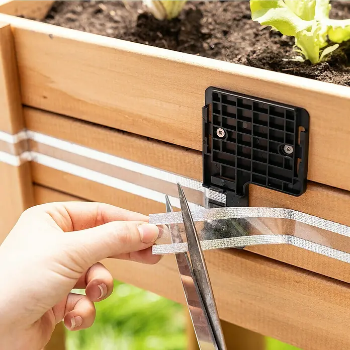 Windhager Schneckenband e-FENCE Tape - 10 m, Passend für: Windhager Schneckenabwehr e-FENCE - Eine Person schneidet mit einer Schere ein silbernes Dichtungsband an einem Holzgewächshaus zu.