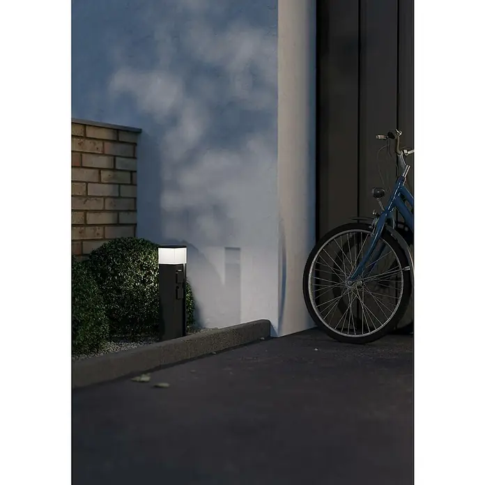Eglo Steckdosensäule Park 5 - Anthrazit, Höhe: 50 cm, IP54 - Eine moderne Außenleuchte in Schwarz beleuchtet einen gepflasterten Weg neben einem Fahrrad und einer Ziegelmauer.