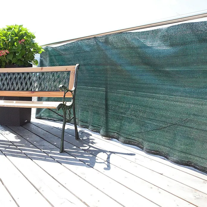 Windhager Schattierungsnetz - 2 x 10 m, Grün - Grüner Sichtschutzzaun auf einer Terrasse mit einer Holzbank und einer Topfpflanze, der Privatsphäre und Windschutz bietet.