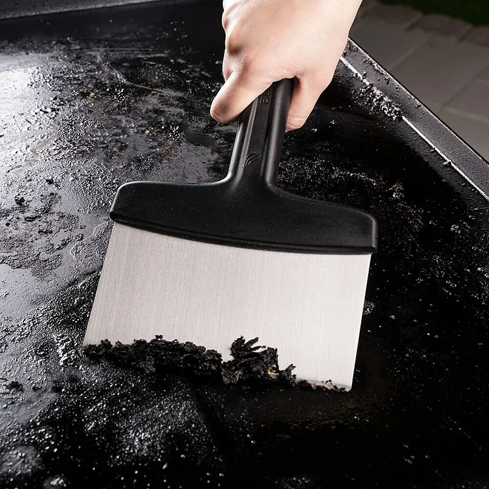 Blackstone Utensilios para barbacoa - Acero inoxidable, 5 pzs. - Una espátula con mango negro limpia una superficie de parrilla negra de residuos de alimentos. Herramienta de cocina práctica para cocinar limpio.