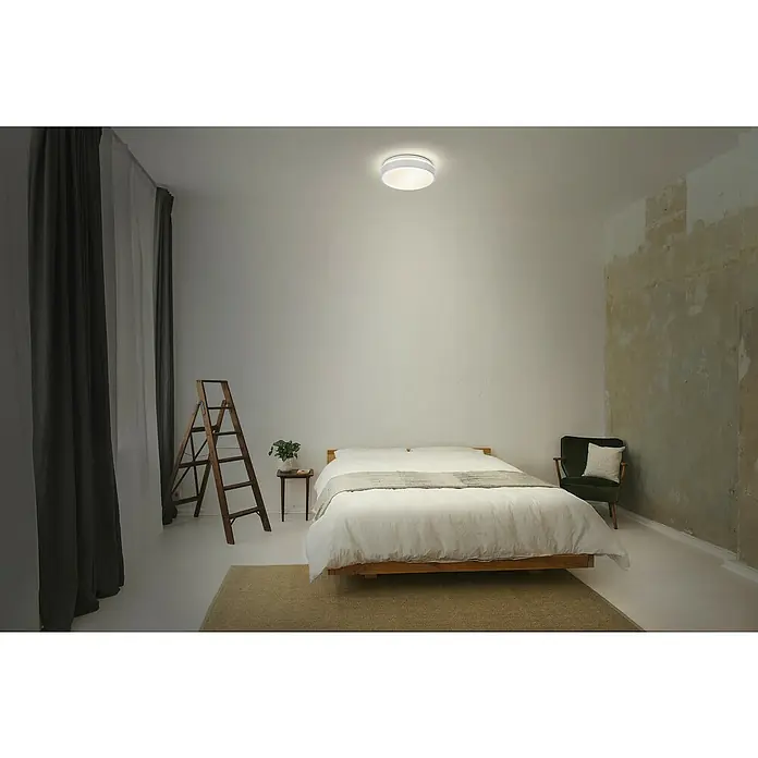 Osram Orbis LED-Deckenleuchte rund Essential - 10 W, Ø x H: 28 x 9,5 cm, Weiß, Ohne Leuchtmittel - Ein modernes Schlafzimmer mit einer runden Deckenleuchte, einem Bett mit weißer Bettwäsche und einem grünen Sessel. Die Leuchte sorgt für angenehmes Licht.