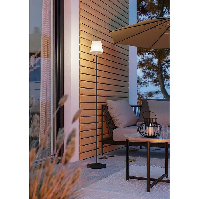 Eglo Solar-Stehleuchte Montapalma-L - Schwarz/ Weiß, IP44, Ø x H: 15 x 140 cm - Eine elegante schwarze Stehlampe mit einem weißen Schirm beleuchtet eine Terrasse mit gemütlichen Möbeln und Holzwänden.