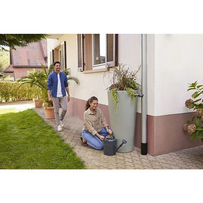 4rain Regenspeicher Vaso Decor 2in1 - 220 l, Agave - Ein schlanker, grauer Regenspeicher mit Bepflanzung, der Regenwasser sammelt. Eine Frau befüllt eine Gießkanne, während ein Mann vorbeigeht.