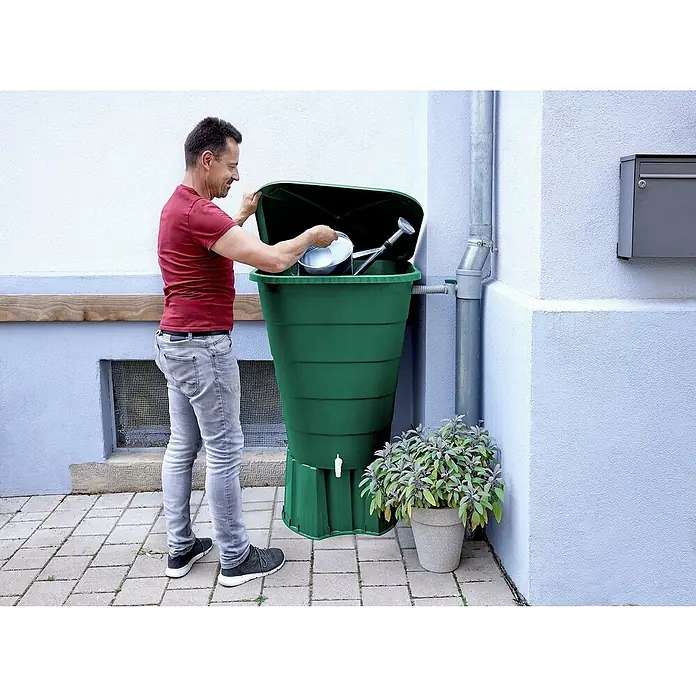 Garantia Regentonne Ibiza - 300 l, Grün, 1 Stk. - Ein Mann befüllt eine grüne Regentonne im Garten. Die Tonne steht neben einem Fallrohr und einem Topf mit Kräutern.