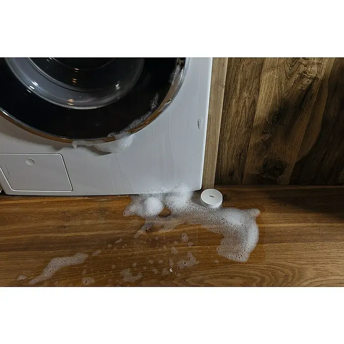 Abus Funk-Wassermelder Smartvest Pro FUWM45000 - Batterielaufzeit: 2 Jahre - Das Bild zeigt einen auslaufenden Waschmaschine mit Schaum auf einem Holzboden, neben einem kleinen, runden, weißen Sensor.