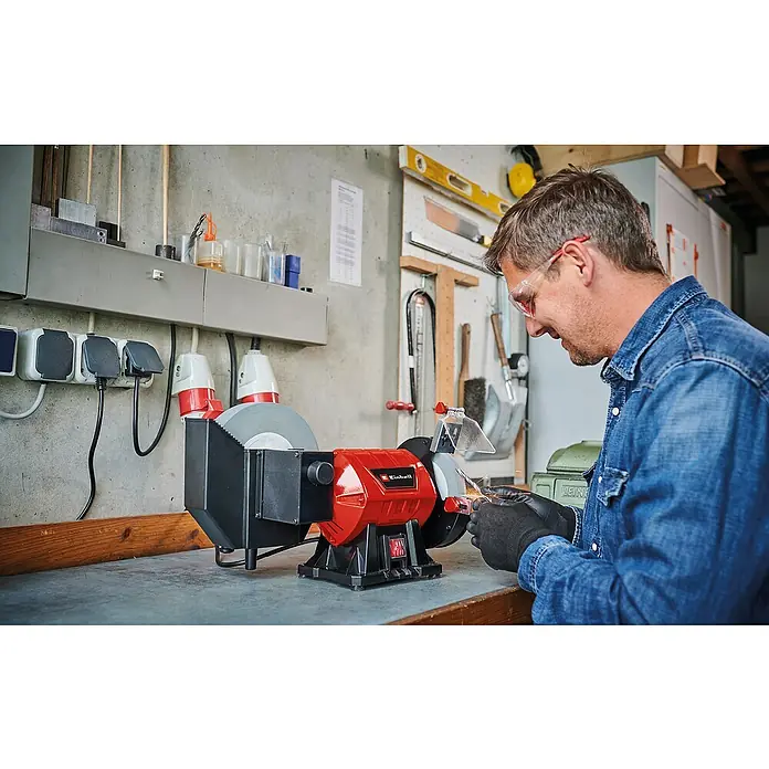 Einhell Nass-Trocken Doppelschleifer TC-WD 200/150 - 170 W, Durchmesser Schleifscheibe: 200 mm, 2.980 U/min - Mann, der eine Einhell Doppelschleifmaschine benutzt, Funken fliegen. Die Maschine steht auf einer Werkbank vor einer Betonwand.